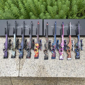 以下が含まれることがあります： 一列に並べられた10個のミニチュア狙撃銃モデルのコレクション。それぞれに稲妻、炎、抽象的なデザインなどのユニークでカラフルな模様が施されています。各モデルにはスコープが装備されており、黒い箱に立てかけられています。