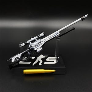 May include: A miniature white and black sniper rifle model featuring intricate graphic patterns and the text "XX" on the side. The rifle is mounted on a black display stand with a white silhouette figure and the letters "CS", accompanied by a gold bullet.