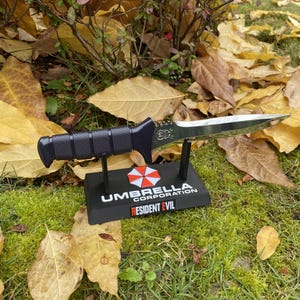 May include: A black and silver knife with a textured handle rests on a black stand. The stand features the text "UMBRELLA CORPORATION" and "RESIDENT EVIL" with a red and white logo. The knife is displayed on a bed of green grass and fallen leaves.