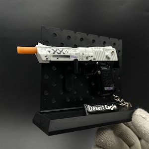 May include: A miniature Desert Eagle toy pistol model featuring a white frame with black geometric patterns, an orange barrel tip, and a black textured grip. The model is mounted on a black pegboard display stand with a label reading Desert Eagle.