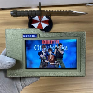 May include: A rectangular, textured gold-colored plastic case featuring a small screen displaying the Resident Evil Code: Veronica video game title and three characters. A blue rectangular label with the word STATUS is above the screen.