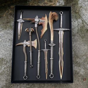 Puede incluir: Una colecci&oacute;n de armas de fantas&iacute;a, incluyendo espadas, hachas y una maza, dispuestas en una bandeja negra. Las armas presentan mangos y hojas plateadas, con detalles color hueso. La bandeja se encuentra sobre un fondo natural al aire libre.