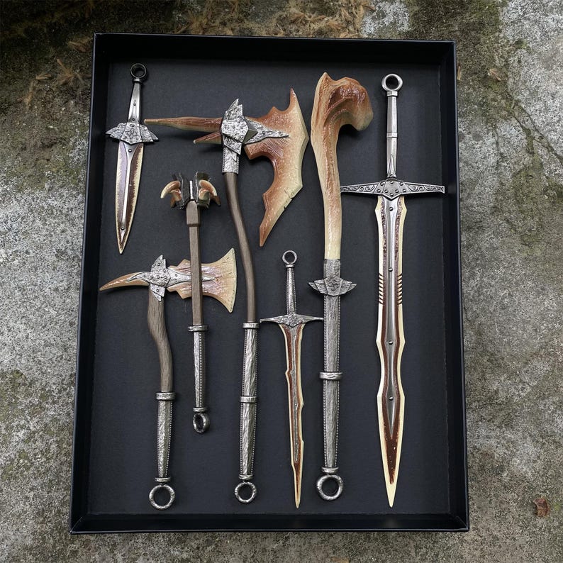 Puede incluir: Una colecci&oacute;n de armas de fantas&iacute;a, incluyendo espadas, hachas y una maza, dispuestas en una bandeja negra. Las armas presentan mangos y hojas plateadas, con detalles color hueso. La bandeja se encuentra sobre un fondo natural al aire libre.