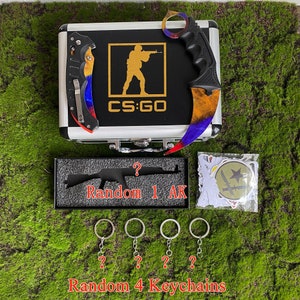 May include: A black case with a CS:GO logo and a gold silhouette of a player holding a gun. The case contains a black knife with a rainbow-colored blade, a black AK-47 keychain, and four silver keychains. The case is sitting on a green surface.