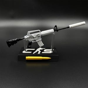 May include: A miniature white and black rifle model with a silencer, mounted on a black display stand featuring the text CS and a silhouette figure. A single yellow bullet replica is positioned in front of the stand on the base.