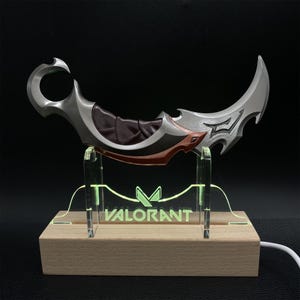 May include: A silver and brown karambit knife with a curved blade, displayed on a wooden base with a green neon "VALORANT" logo. The knife has a dark brown handle and red accents.