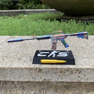 May include: A miniature rifle model featuring a colourful graffiti-style wrap with the word DANGER printed on the side. The model is mounted on a black rectangular stand with the letters CS and a white silhouette figure, plus a gold bullet replica.
