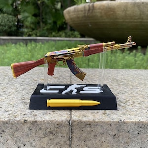 May include: A miniature rifle model featuring a gold-toned receiver, a reddish-brown stock and grip, and a multi-colored magazine. The model is mounted on a black rectangular stand with white lettering and a gold-colored bullet replica.