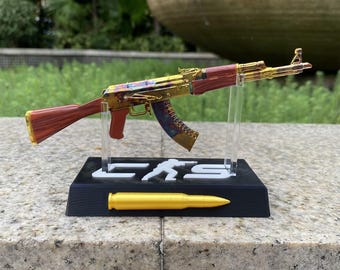 May include: A miniature rifle model featuring a gold-toned receiver, a reddish-brown stock and grip, and a multi-colored magazine. The model is mounted on a black rectangular stand with white lettering and a gold-colored bullet replica.