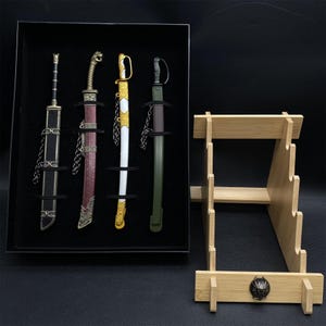 May include: A set of four decorative miniature swords in a black display box, each with unique handle and sheath designs. A wooden sword stand is next to the box. The swords have gold, silver, black, white, and burgundy accents.