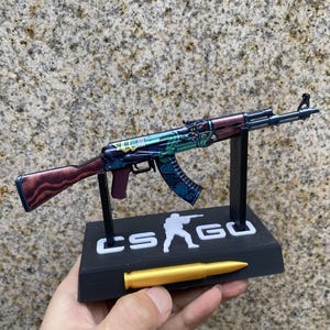 May include: A miniature AK-47 rifle replica with a colorful, patterned design. The rifle has a wooden stock and is displayed on a black stand with the text "CS:GO". A gold-colored bullet is placed in front.