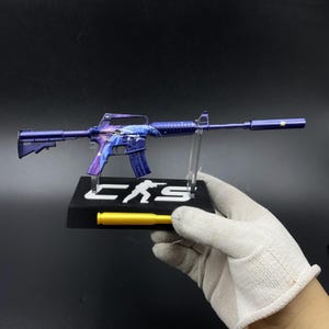 May include: A miniature purple rifle model with a galaxy-themed finish, featuring a silencer and a carry handle. The model is mounted on a black display stand with the white text CS and a silhouette figure, plus a yellow bullet-shaped accent.