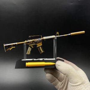 May include: A miniature model of an assault rifle featuring a black and gold marbled pattern finish. The rifle is mounted on a clear plastic stand attached to a black base, which includes a single gold-coloured bullet replica attached to the front.