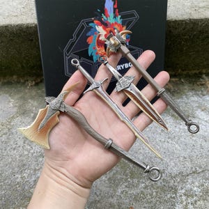 Puede incluir: Cuatro armas de fantas&iacute;a en miniatura: un hacha, dos espadas y una maza. El hacha tiene un mango de madera y una hoja dorada y plateada. Las espadas y la maza tienen hojas y empu&ntilde;aduras plateadas. Las armas se sostienen en una mano.