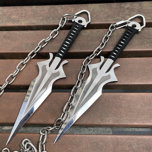 Può includere: Due lame kunai in metallo argentato con punte acuminate e bordi taglienti. Ogni lama presenta un manico avvolto da un cordino nero e un anello in metallo sul pomolo. Una lunga e pesante catena in metallo argentato collega le due armi.