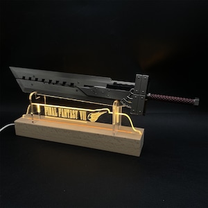 May include: A detailed replica of a Buster Sword from Final Fantasy VII, resting on a wooden base with the game's logo illuminated in yellow. The sword is metallic gray with a brown handle, and the base has a warm, inviting glow.