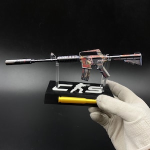 May include: A miniature rifle model featuring a colourful graphic print on the receiver and magazine. The model is mounted on a black display stand with the white text "CS" and a silhouette icon, accompanied by a yellow cylindrical component.
