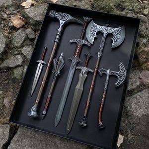 May include: A collection of fantasy weapons, including swords, axes, and a mace, arranged in a black tray. The weapons feature silver and bronze tones with intricate designs, suggesting a medieval or fantasy theme.