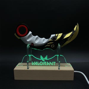 May include: A curved, decorative knife with a black, gold, and silver design, displayed on a clear stand with the word "VALORANT" illuminated in green. The base is made of light-colored wood.