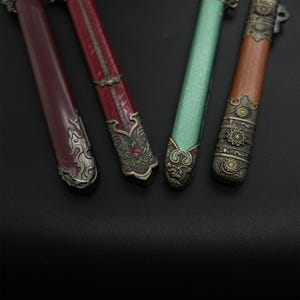 May include: Close-up of four decorative sword scabbards. The scabbards are in shades of red, green, and brown, with ornate metal detailing. The metalwork features intricate patterns and embellishments, including floral and geometric designs.