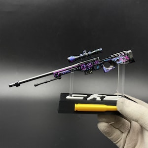 May include: A miniature sniper rifle model featuring a black barrel and a purple and blue graffiti-patterned stock with white text. The rifle includes a scope, a bipod, and a black display stand with a yellow bullet-shaped accent piece.