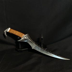 May include: A silver and brown dagger with a curved blade and a serrated edge. The handle is wrapped in brown leather. The hilt features decorative silver accents. The dagger is displayed on a black stand against a black background.