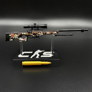 Könnte beinhalten: Ein Miniatur-Scharfschützengewehr-Modell mit schwarzem Lauf und Zielfernrohr sowie einem bunten Cartoon-Figuren-Muster auf dem Schaft. Das Gewehr ist auf einem schwarzen Ständer mit dem weißen Text CS und einer Silhouette montiert, begleitet von einer goldenen Patrone.