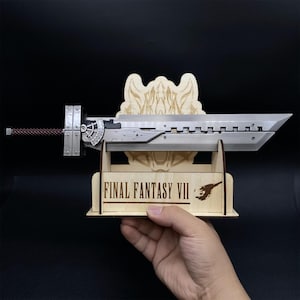 May include: A silver-colored Buster Sword replica from Final Fantasy VII, displayed on a wooden stand. The stand features the game's logo and a stylized design. The sword has a detailed hilt and a textured handle.