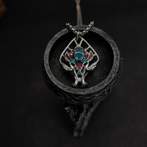 Pode incluir: Um colar com um pingente de prata com um design detalhado que apresenta uma flor estilizada com detalhes em esmalte vermelho, verde e azul. O pingente está pendurado em uma corrente de prata.