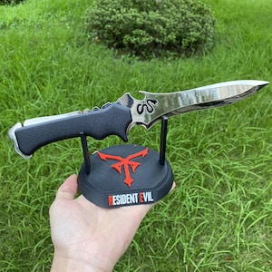 May include: A silver and black dagger with a serrated spine, resting on a black stand. The stand features a red emblem and the text "RESIDENT EVIL". The dagger has a curved blade and a black handle.