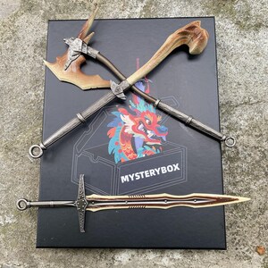 Puede incluir: Un surtido de armas de fantas&iacute;a, que incluyen un hacha de batalla, una espada y una caja con el texto "MYSTERYBOX". El hacha presenta una hoja marr&oacute;n y un mango similar a un hueso. La espada tiene una hoja marr&oacute;n con un mango plateado. La caja tiene una ilustraci&oacute;n de drag&oacute;n colorido.