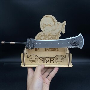 Può includere: Un espositore in legno con una spada nera e un personaggio del videogioco Nier. L'espositore ha la parola "Nier" incisa.