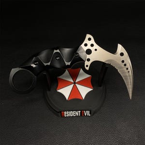 Op de afbeelding: Een karambitmes met een zwart handvat en een zilverkleurig lemmet. Het mes staat op een zwarte standaard met het Resident Evil-logo. Het lemmet heeft een gebogen ontwerp met meerdere gaten en een gekartelde rand.
