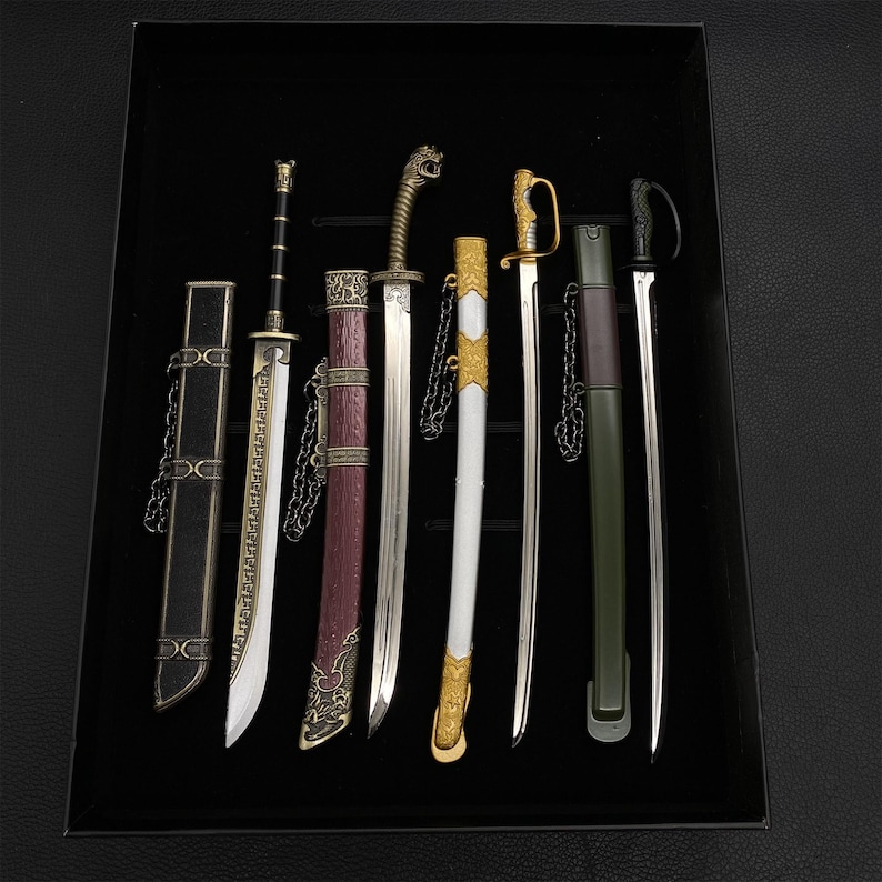 May include: A collection of miniature swords with ornate handles and sheaths, displayed in a black box. The swords vary in design, with silver blades and decorative elements in gold, black, and burgundy. Some have chains attached to the sheaths.