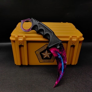 Op de afbeelding: Een karambitmes met een zwart handvat en een gebogen lemmet met een levendig melkwegstelselontwerp in paars, blauw en roze. Het mes heeft een vingerring en staat op een gele container.