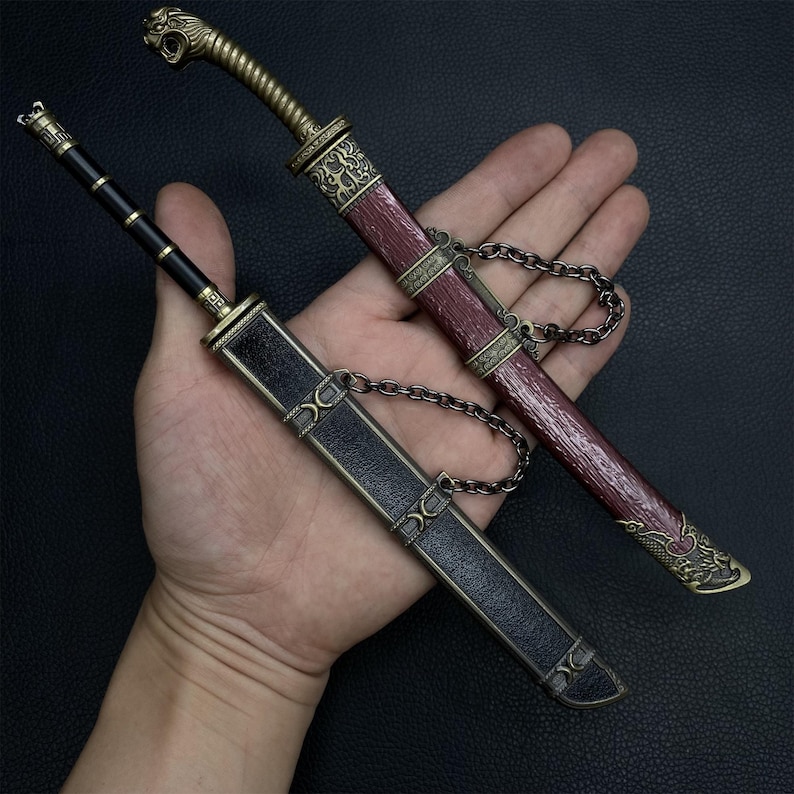 May include: Two decorative miniature swords with sheaths. One sword has a black handle and sheath, the other has a red handle and sheath. Both have ornate gold-colored metal accents and are connected to their sheaths by a chain.
