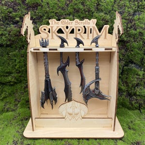 May include: A wooden display shelf featuring the text "The Elder Scrolls V SKYRIM" and various miniature weapons. The shelf is made of light-colored wood and has a decorative dragon design on the sides.