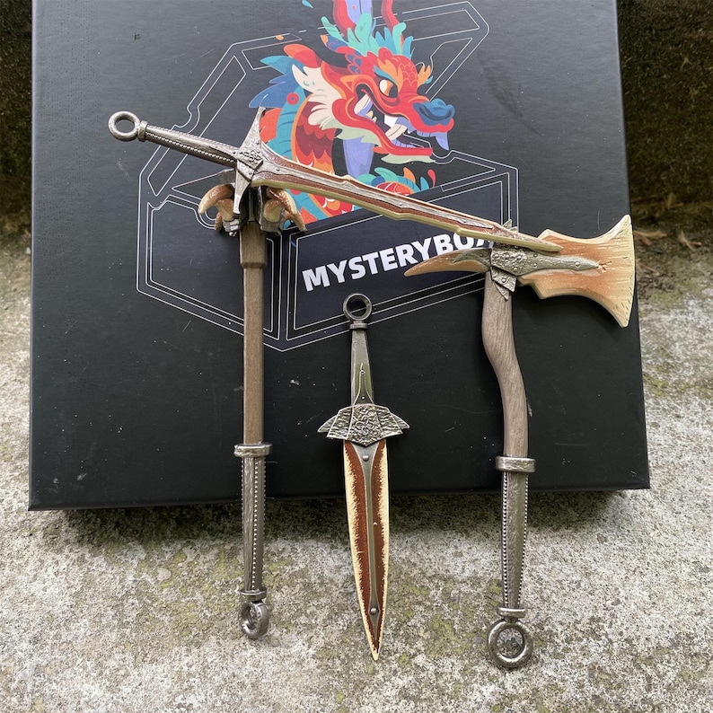 Puede incluir: Tres armas de fantas&iacute;a en miniatura: una espada, una daga y un hacha. La espada tiene una hoja y un mango plateados, la daga tiene una hoja marr&oacute;n y plateada, y el hacha tiene un mango de madera y una cabeza plateada. Las armas se exhiben frente a una caja con el texto "MYSTERYBOX".