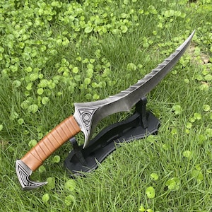 May include: A decorative dagger with a serrated blade and a brown leather-wrapped handle. The dagger has silver-colored accents with geometric designs and is displayed on a black stand, set against a backdrop of green grass.