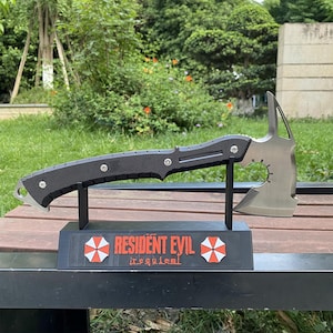 May include: A black and silver axe with a textured handle and a tomahawk-style head. The axe is displayed on a black stand with the text "RESIDENT EVIL" and a red and white umbrella logo.