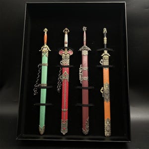 May include: Four miniature decorative swords displayed in a black case. The swords have ornate handles and sheaths in green, red, and orange. Each sword is secured by a black strap. The swords feature decorative metal accents and chain details.
