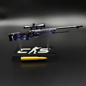 May include: A miniature sniper rifle model featuring a purple and blue marbled pattern, equipped with a scope and bipod. The rifle rests on a black display stand with the white text "CS" and a silhouette figure, accompanied by a single yellow bullet.