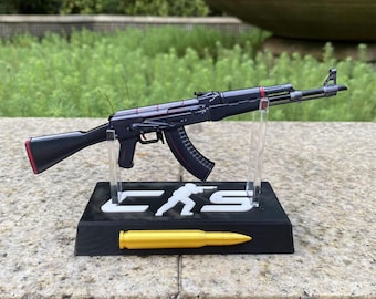 May include: A miniature black rifle model with red accents mounted on a black display stand. The stand features the white text CS with a silhouette of a person holding a rifle and a gold-colored bullet replica attached to the front base.