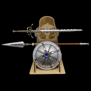 Pode incluir: Exibição de armas de fantasia, incluindo uma espada prateada com punho decorativo, uma lança e um escudo prateado com um centro de gema azul. Os itens são exibidos em um suporte de madeira com um design de caveira.