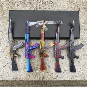 Puede incluir: Cinco modelos de rifles en miniatura dispuestos en fila frente a una caja rectangular negra. Los rifles presentan acabados distintos que incluyen oro metálico, degradado azul y morado, veta de madera, rojo y negro mate, con piezas mecánicas detalladas.
