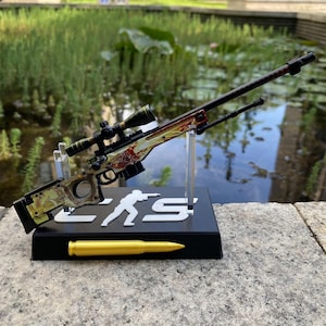 May include: A miniature sniper rifle model featuring a detailed gold and red patterned stock, a black scope, and a bipod. The model is mounted on a black display stand with a white silhouette logo and a gold-colored bullet replica at the base.
