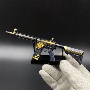 May include: A miniature rifle model featuring a dark blue and gold finish with geometric patterns, including diamond shapes on the magazine and receiver. The toy firearm is mounted on a black display stand, showcasing intricate metallic detailing.