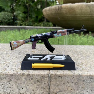 May include: A miniature black rifle model featuring a brown stock decorated with various colorful stickers, mounted on a black display stand with the text CS and a silhouette figure, featuring a gold-colored bullet replica on the front base.