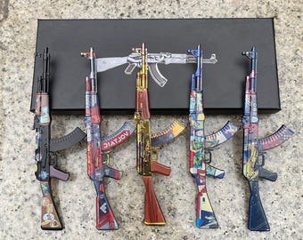 May include: Five miniature rifle models arranged in a row on a black rectangular box. Each model features a distinct finish, including solid black, red and blue patterns, metallic gold, multi-colored geometric designs, and dark blue.