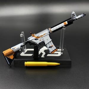 May include: A miniature rifle model featuring a white, black, and orange geometric paint scheme, mounted on a black display stand with a white silhouette logo. A single yellow bullet-shaped accessory is positioned in front of the stand.